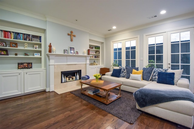 living room with crown molding, dark hardwood / wood-style floors, a tiled fireplace, built in shelves, and french doors