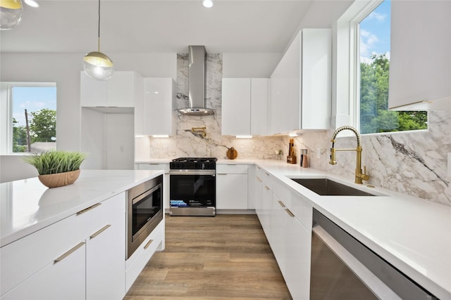 kitchen with sink, hanging light fixtures, stainless steel appliances, white cabinets, and wall chimney exhaust hood