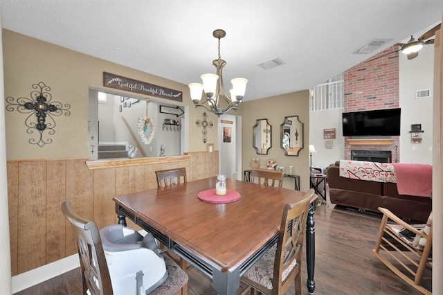 dining room with dark wood-type flooring, wooden walls, a brick fireplace, and a notable chandelier