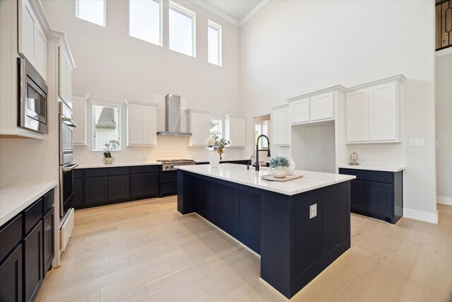 kitchen with white cabinetry, appliances with stainless steel finishes, a center island with sink, and wall chimney range hood