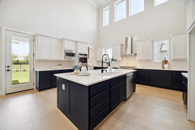 kitchen with sink, white cabinets, wall chimney range hood, stainless steel appliances, and a center island with sink