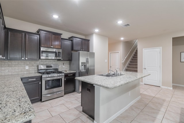 kitchen with appliances with stainless steel finishes, sink, decorative backsplash, a kitchen island with sink, and light stone countertops