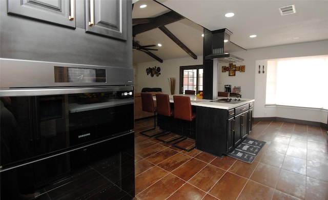 kitchen featuring lofted ceiling with beams, oven, a kitchen bar, ceiling fan, and wall chimney range hood