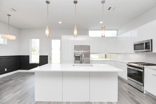 kitchen with appliances with stainless steel finishes, a kitchen island with sink, backsplash, white cabinets, and decorative light fixtures