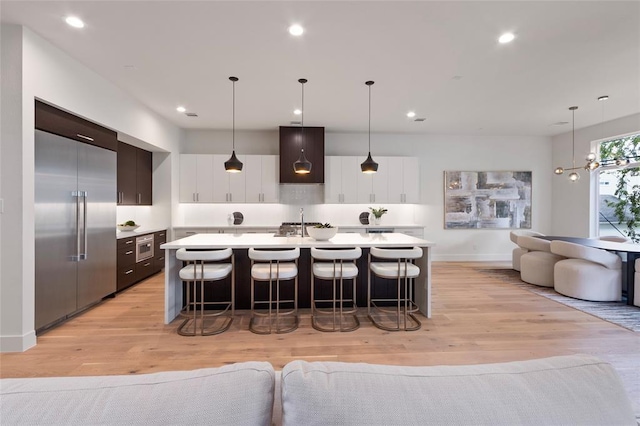 kitchen featuring white cabinetry, dark brown cabinetry, an island with sink, decorative light fixtures, and stainless steel built in fridge