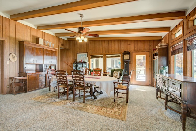 dining room featuring light carpet, wood walls, beamed ceiling, and a healthy amount of sunlight