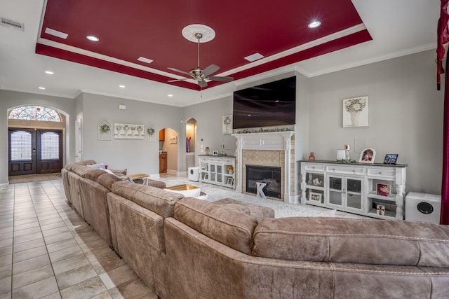 living room with french doors, ornamental molding, a tray ceiling, a tile fireplace, and ceiling fan