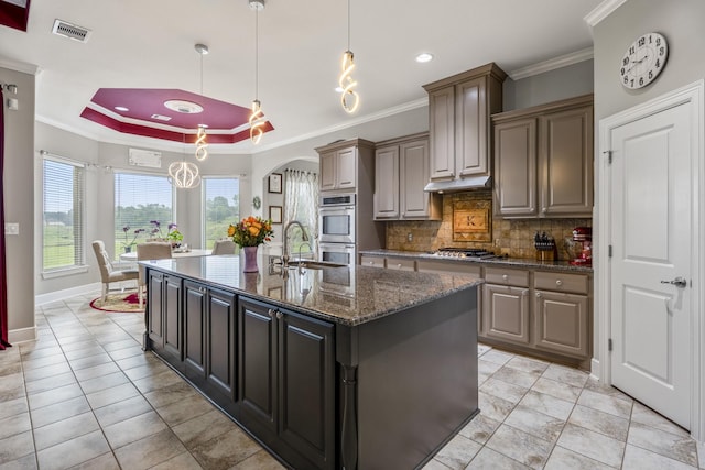 kitchen featuring appliances with stainless steel finishes, pendant lighting, a raised ceiling, crown molding, and a center island with sink