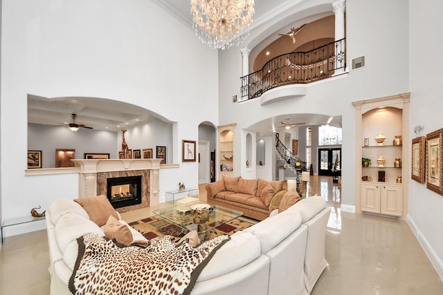 living room with crown molding, ceiling fan with notable chandelier, a towering ceiling, a high end fireplace, and built in shelves