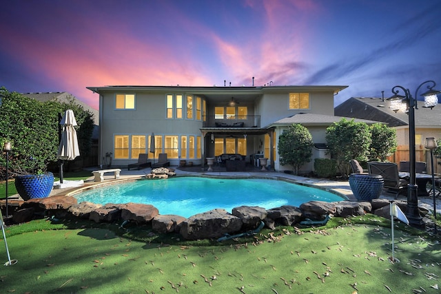 back house at dusk featuring a patio, a balcony, ceiling fan, and a lawn