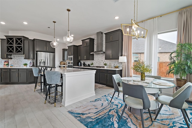 kitchen featuring pendant lighting, backsplash, wall chimney range hood, stainless steel appliances, and a center island with sink