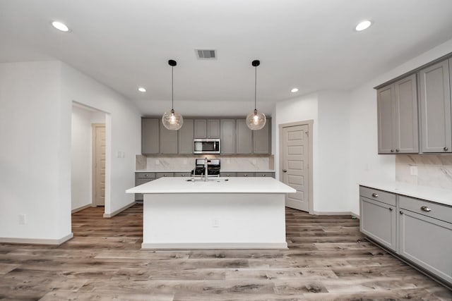 kitchen with dark wood-type flooring, tasteful backsplash, appliances with stainless steel finishes, gray cabinets, and an island with sink