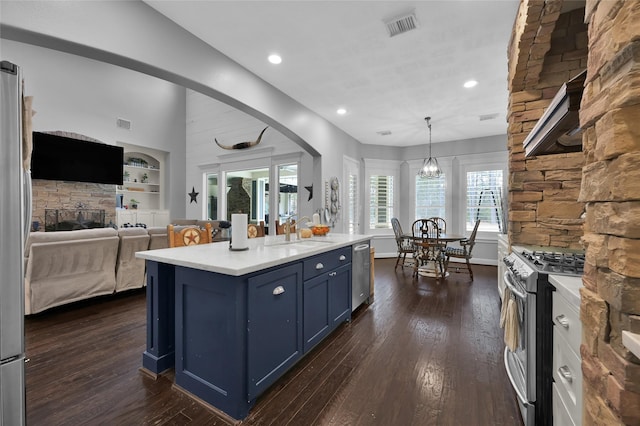 kitchen featuring appliances with stainless steel finishes, blue cabinets, hanging light fixtures, a kitchen island with sink, and dark wood-type flooring