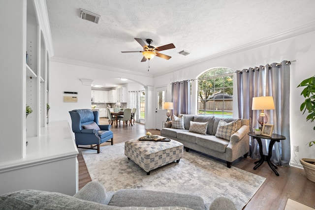 living area with arched walkways, wood finished floors, visible vents, decorative columns, and crown molding