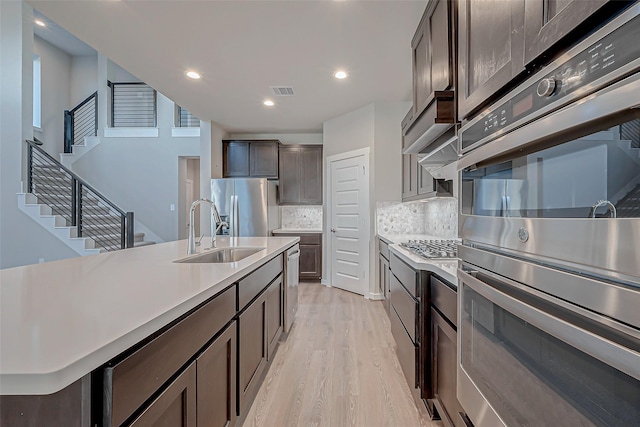 kitchen with sink, tasteful backsplash, a center island with sink, light wood-type flooring, and appliances with stainless steel finishes