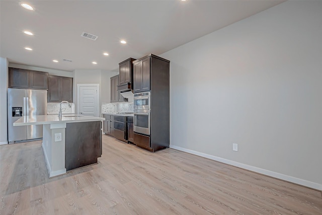 kitchen featuring sink, light hardwood / wood-style flooring, appliances with stainless steel finishes, an island with sink, and backsplash