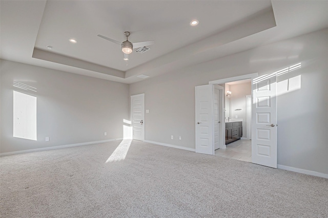 unfurnished bedroom with light carpet, a tray ceiling, ensuite bath, and ceiling fan