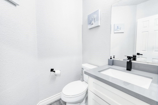 half bath with toilet, a textured wall, baseboards, and vanity