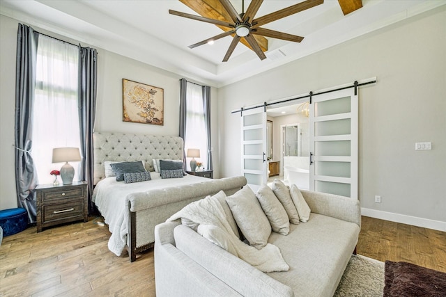 bedroom with ceiling fan, connected bathroom, a tray ceiling, light hardwood / wood-style floors, and a barn door