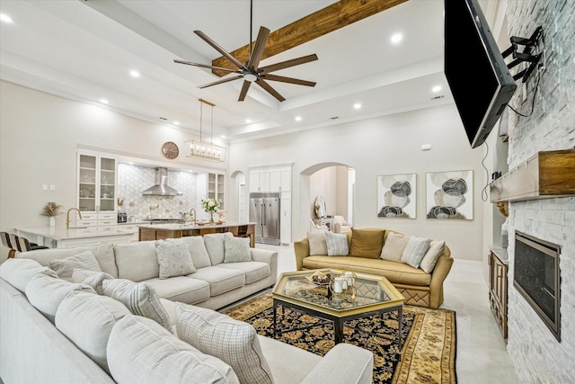 living room featuring a stone fireplace, sink, ceiling fan, beam ceiling, and a high ceiling