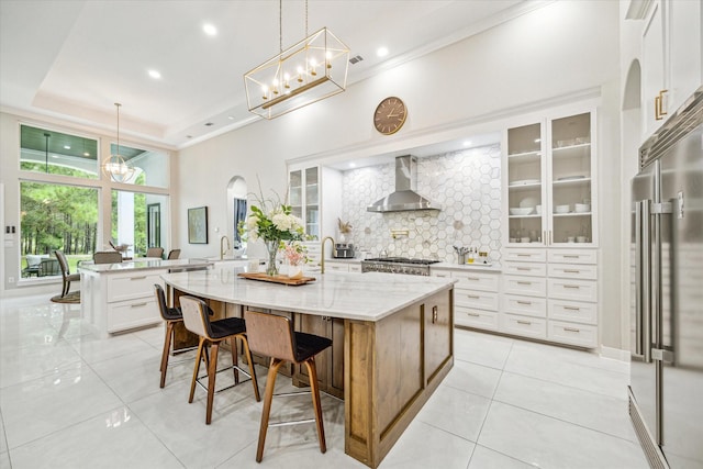 kitchen with wall chimney range hood, light stone counters, an island with sink, white cabinets, and high end refrigerator