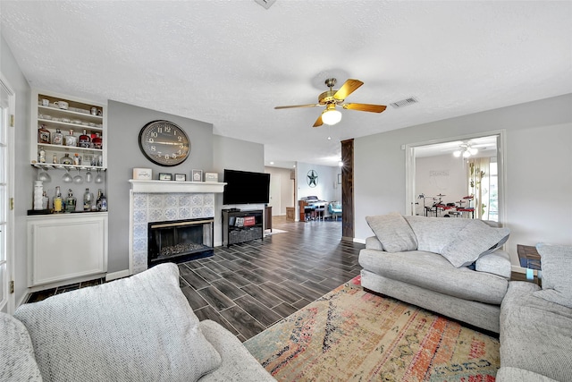 living room featuring built in shelves, ceiling fan, a tiled fireplace, and a textured ceiling