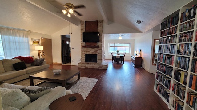 living room with dark wood-type flooring, lofted ceiling with beams, a brick fireplace, a textured ceiling, and ceiling fan