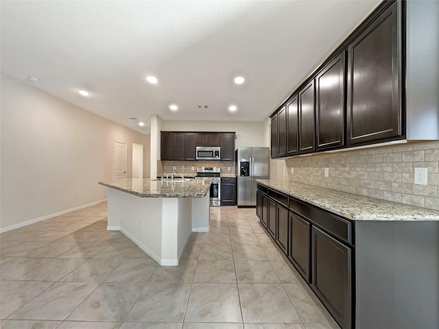 kitchen featuring light tile patterned floors, stainless steel appliances, light stone counters, an island with sink, and decorative backsplash