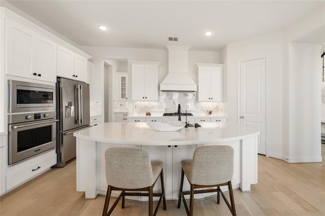 kitchen featuring appliances with stainless steel finishes, backsplash, an island with sink, custom range hood, and white cabinets