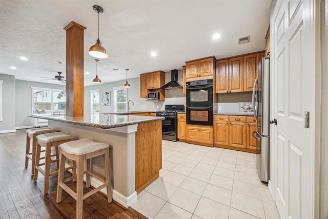 kitchen featuring a kitchen bar, black appliances, dark stone countertops, pendant lighting, and wall chimney range hood