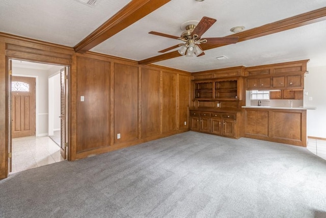 unfurnished living room featuring crown molding, ceiling fan, beam ceiling, wooden walls, and light colored carpet