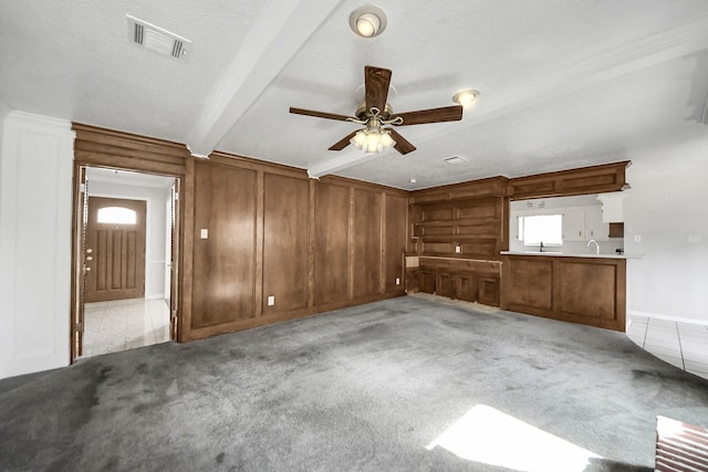 unfurnished living room featuring wood walls, a textured ceiling, ceiling fan, beamed ceiling, and light colored carpet