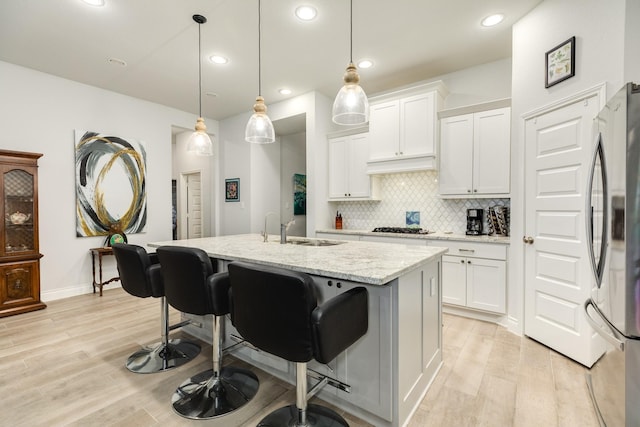 kitchen with stainless steel refrigerator, a kitchen island with sink, hanging light fixtures, light stone counters, and white cabinets