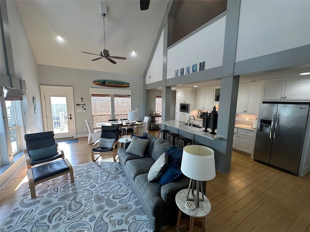 living room with ceiling fan, high vaulted ceiling, sink, and light wood-type flooring