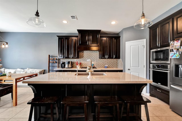 kitchen with dark brown cabinetry, light stone counters, light tile patterned floors, stainless steel appliances, and a kitchen island with sink