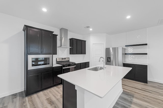 kitchen featuring wall chimney exhaust hood, sink, an island with sink, stainless steel appliances, and light hardwood / wood-style floors