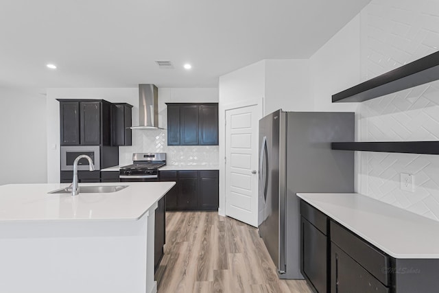 kitchen with wall chimney range hood, sink, appliances with stainless steel finishes, a kitchen island with sink, and light wood-type flooring