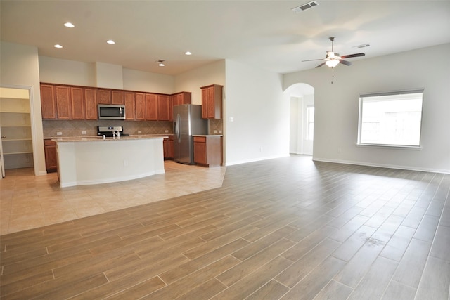 kitchen with appliances with stainless steel finishes, backsplash, ceiling fan, a center island with sink, and light hardwood / wood-style flooring