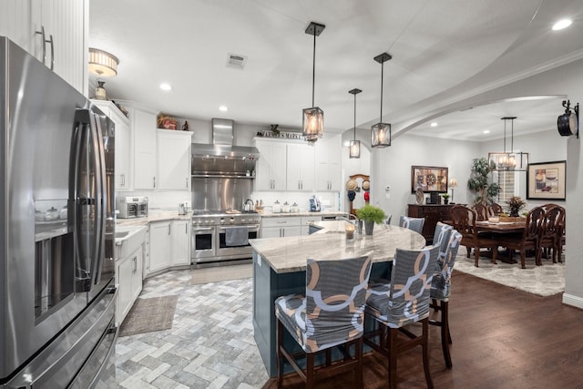 kitchen featuring decorative light fixtures, wall chimney range hood, stainless steel appliances, a kitchen island with sink, and white cabinets