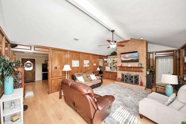 living room with ceiling fan, vaulted ceiling with beams, wooden walls, a brick fireplace, and light wood-type flooring