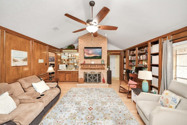 living room featuring vaulted ceiling, a fireplace, wooden walls, ceiling fan, and light wood-type flooring