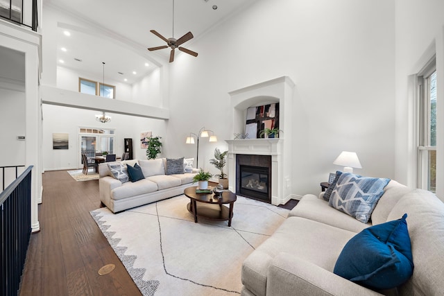 living room featuring a tiled fireplace, wood-type flooring, a high ceiling, and ceiling fan with notable chandelier