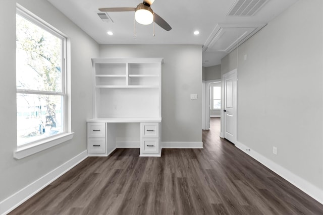 unfurnished office featuring ceiling fan, a wealth of natural light, built in desk, and dark wood-type flooring