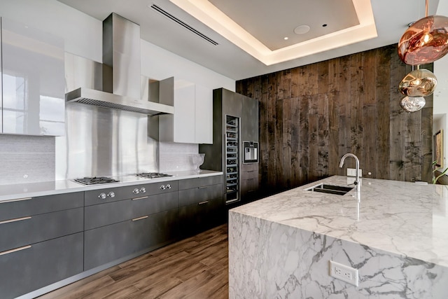 kitchen featuring wood walls, sink, white cabinets, a tray ceiling, and wall chimney exhaust hood