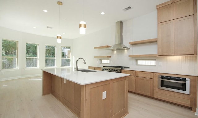 kitchen with pendant lighting, sink, a kitchen island with sink, stainless steel appliances, and wall chimney exhaust hood