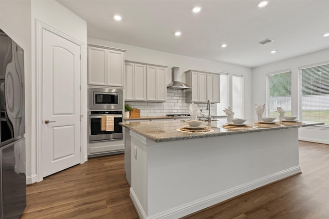 kitchen with a center island with sink, appliances with stainless steel finishes, wall chimney range hood, light stone countertops, and white cabinets