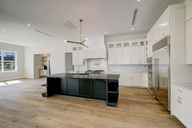 kitchen featuring premium range hood, backsplash, a kitchen island with sink, stainless steel built in fridge, and light hardwood / wood-style flooring