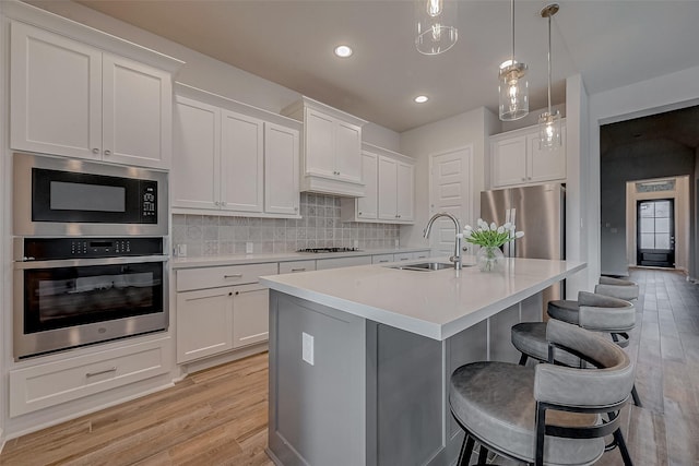 kitchen featuring a kitchen island with sink, light wood-style flooring, a sink, appliances with stainless steel finishes, and decorative backsplash