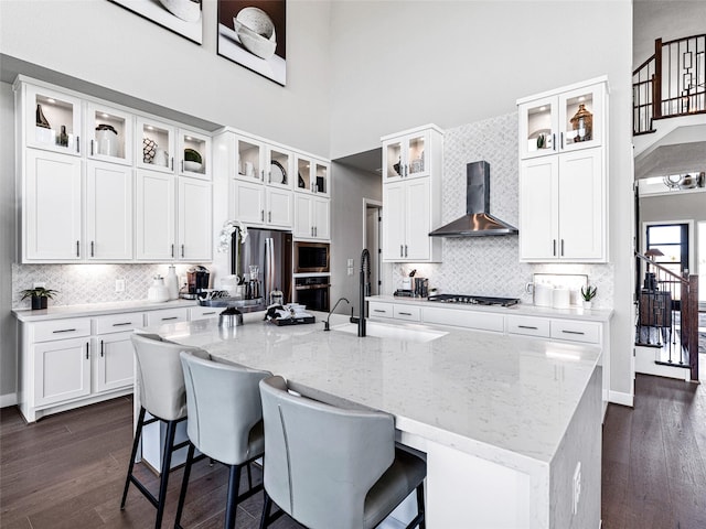 kitchen with light stone counters, an island with sink, wall chimney exhaust hood, and appliances with stainless steel finishes