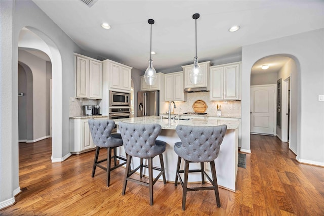 kitchen featuring decorative light fixtures, an island with sink, stainless steel appliances, light stone countertops, and white cabinets